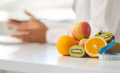 Close-up of pile of fresh apples, kiwis and oranges and measuring tape on office desk against blurred dietitian or nutritionist giving weight loss advice during consultation with patient